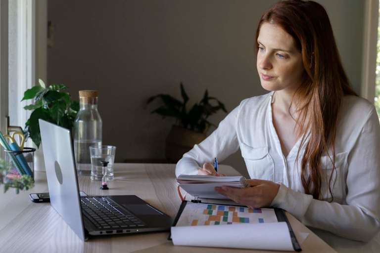 Psychologist writing notes in a book while working on her laptop at home.